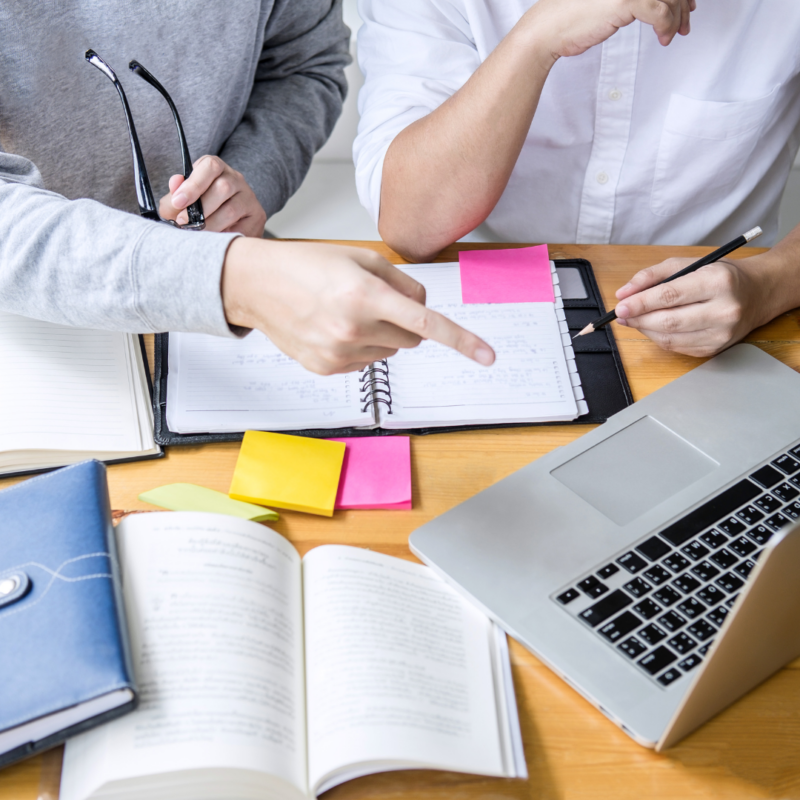 Chest and arms of two people are visible. Person on the left is pointing at a laptop screen. Books and a laptop are on the table in front of them.